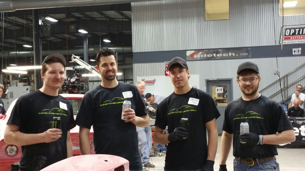 Four men in black shirts standing next to a car in a garage.