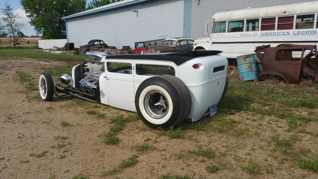 A white hot rod is parked in a dirt lot.